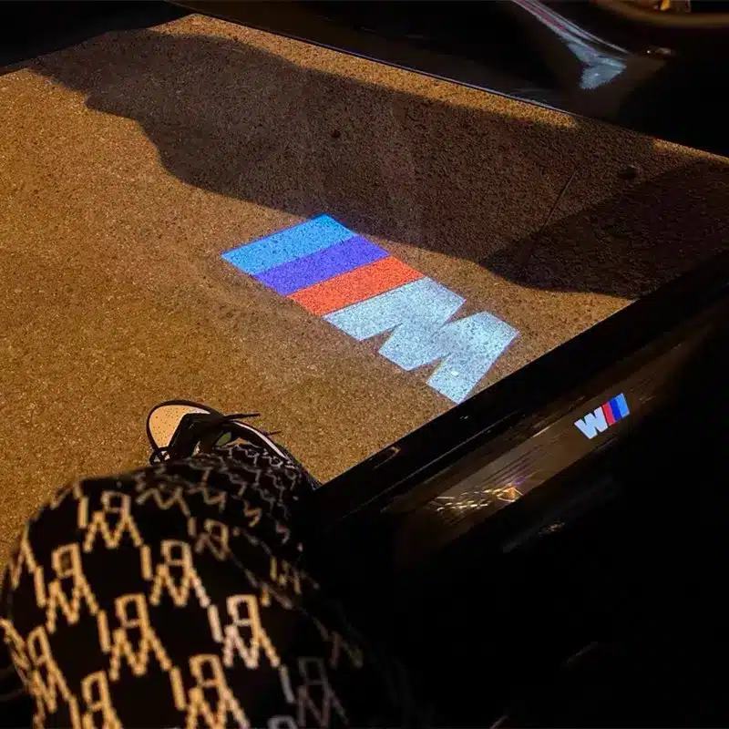 BMW M Door Lights| LED BMW M Door Lights Car door open showing an illuminated BMW M logo projected on the ground. Person wearing patterned pants and sneakers partially visible, creating a modern and stylish vibe.