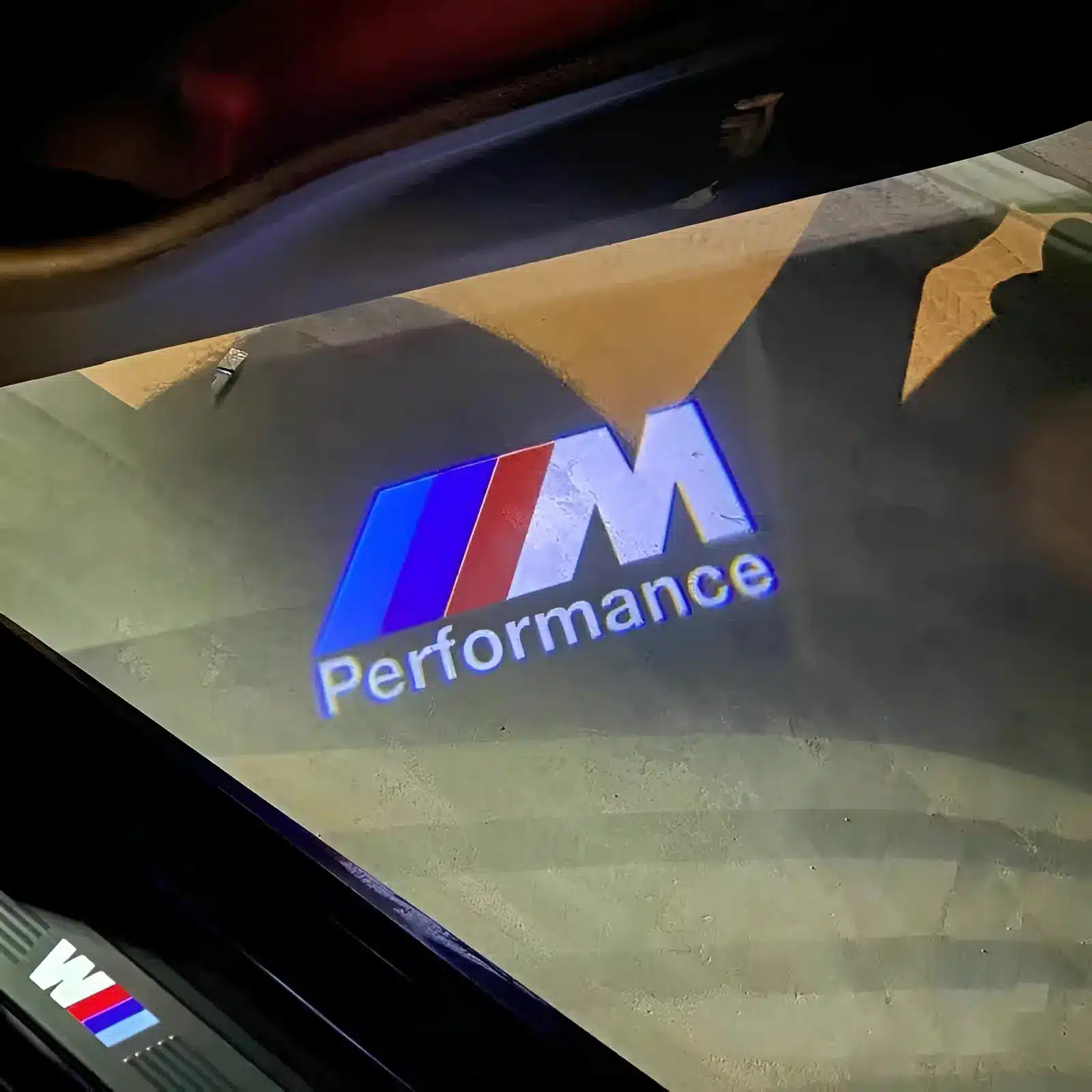 BMW M Sport Door Lights| BMW M Sport Door Lights Logo Projector Floor illuminated with a "M Performance" logo featuring blue, purple, and red stripes, under a car door. The image conveys a sleek, modern vibe.